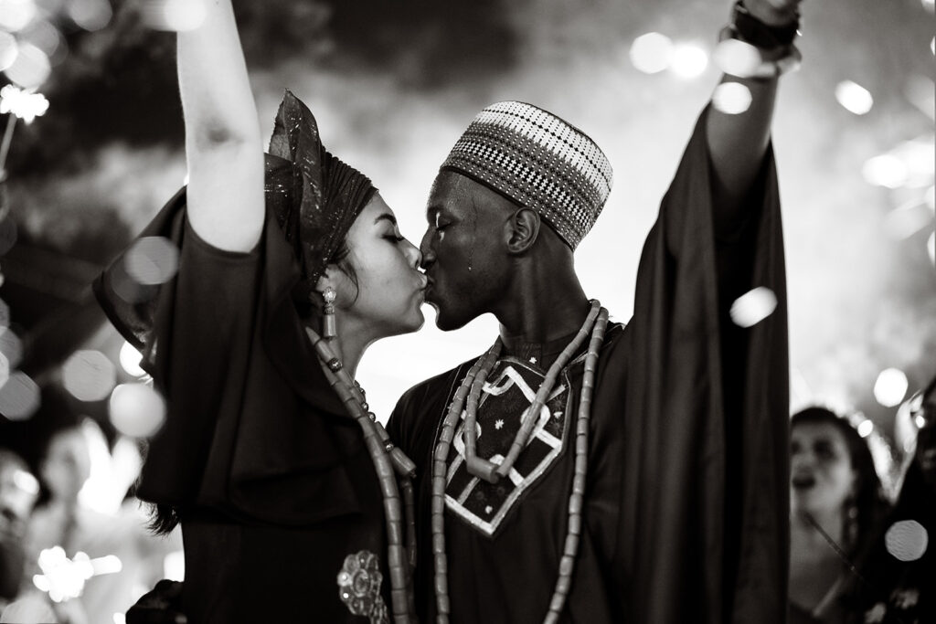 A couple in Nigerian wedding garb kisses while their friends and family cheer for them holding sparklers at their Roatan wedding.  