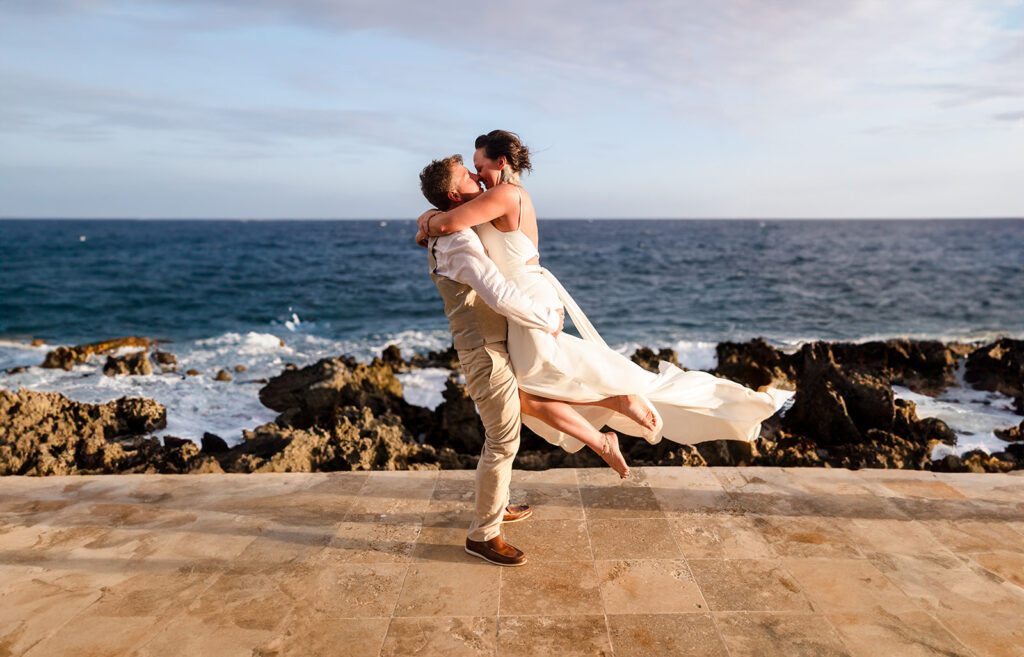 A groom picks up his bride and they kiss while waves crash on the Iron Shore in Roatan, Honduras