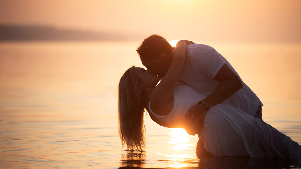 A groom dips his bride and kisses her while they stand in the ocean on their casual wedding day