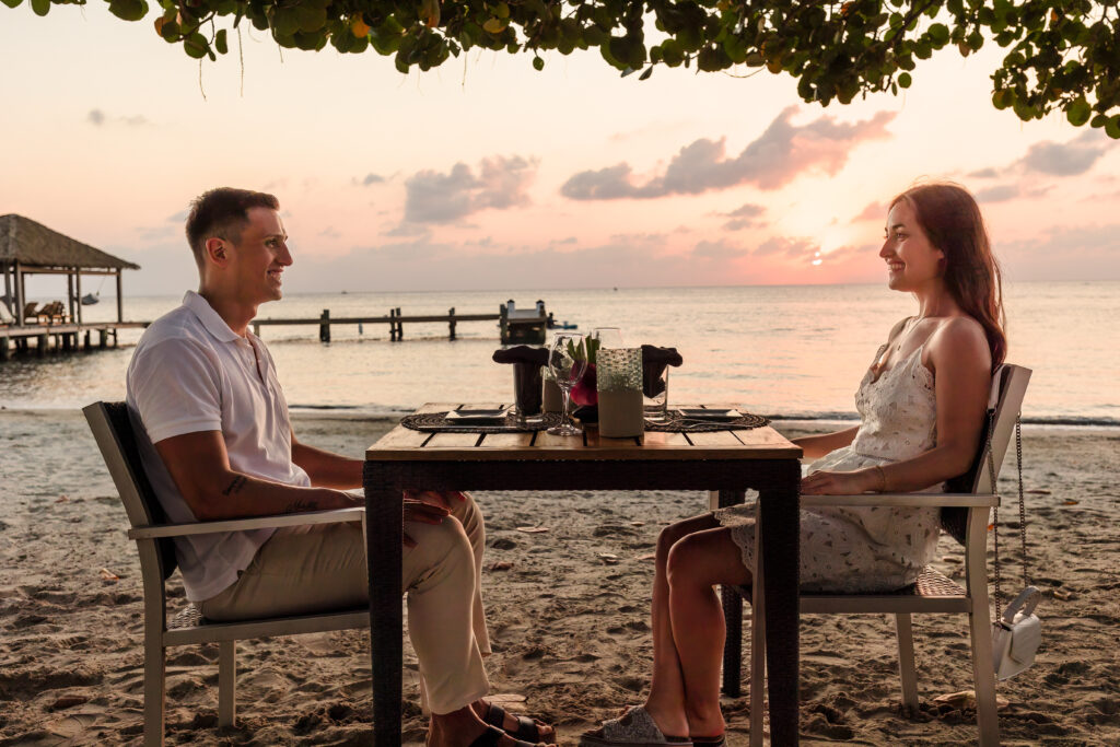 A couple sit at a table on the beach at sunset smiling at eachother.