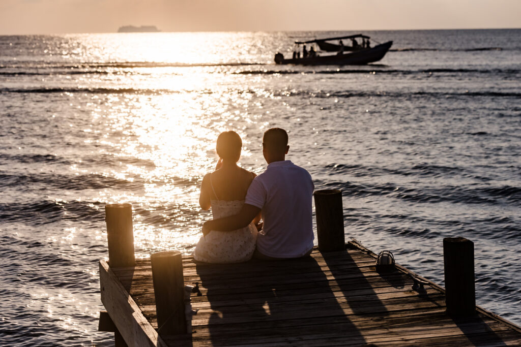 A couple sit on a dock watching a sunset as a boat goes by.