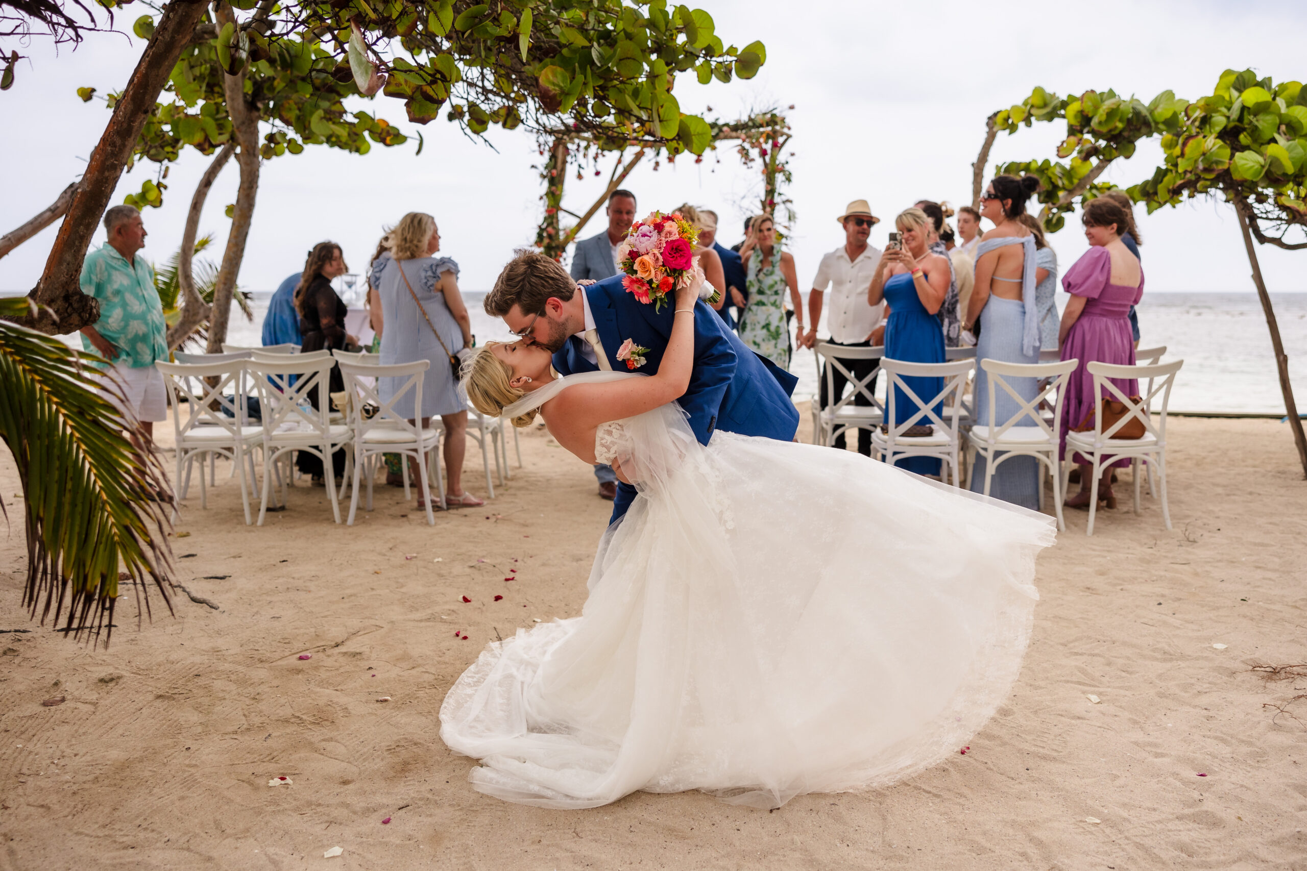 A groom dips and kisses his bride on the beach in front of guests.