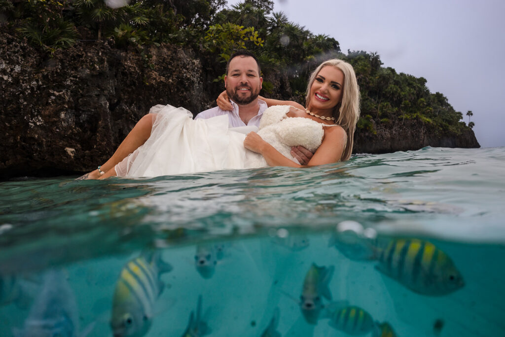 A couple in the ocean at West Bay Beach in Roatan during their trash the dress photo session. The photo is a split shot between above and below the water with small tropical fish in the foreground of the photo. 