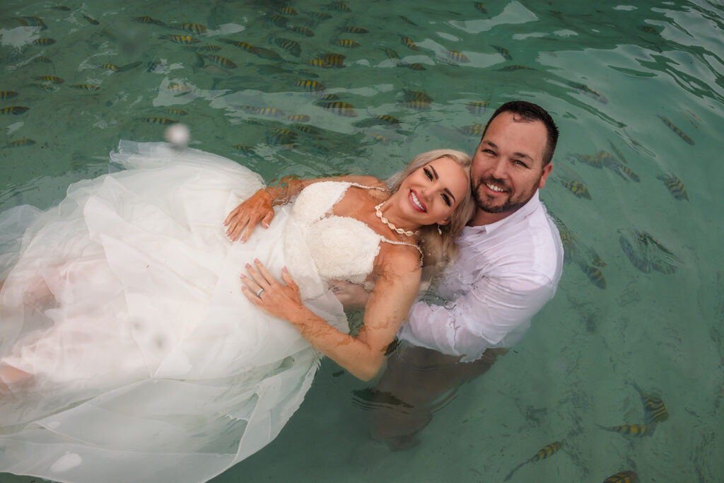 A bride and groom lay in the sea in her wedding dress in the ocean at West Bay Beach in Roatan during their trash the dress photo session. 