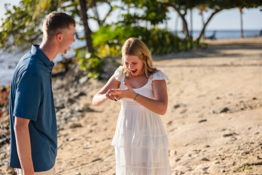 A woman smiles and admires her ring while her fiance watches her right after their beach proposal in Roatan.