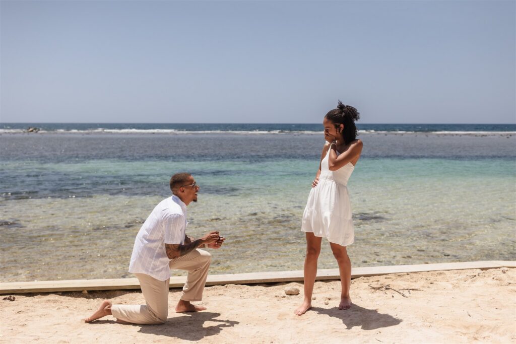 A man is on one knee in front of a white sand beach in Roatan proposing to his girlfriend and she is shocked and happy.