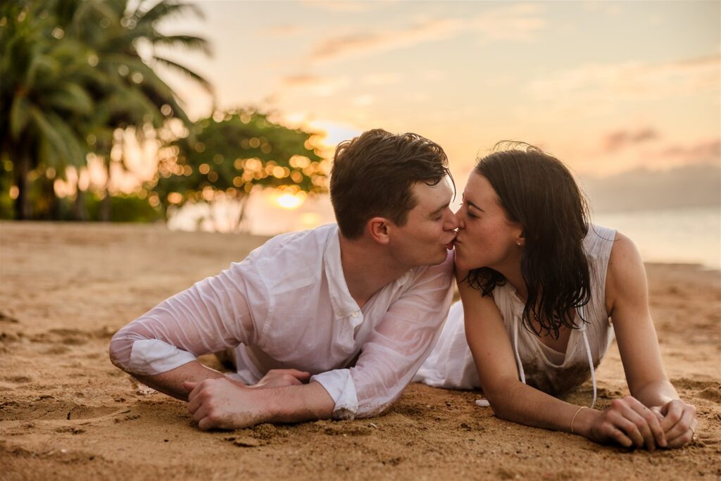 A couple kisses while lying on their stomachs in the sand in Roatan after their Roatan proposal.